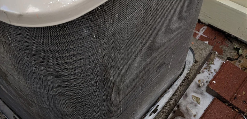 Close-up of an outdoor air conditioning unit being cleaned, with soap suds and water visible on the concrete base and nearby bricks.