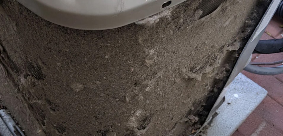 A close-up of an air conditioner coil covered in a thick layer of dust, dirt, and debris, indicating poor maintenance and restricted airflow.