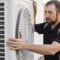 Professional HVAC Installation and Repair A man with a beard inspects or installs an outdoor air conditioning unit, placing his hands on the side of the machine. He appears to be focused on his work.